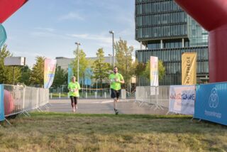 Zwei Läufer im grünen Shirt laufen gemeinsam ins Ziel beim Förderturm-Firmenlauf – Start-Ziel-Bogen und Banner im Hintergrund