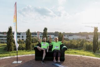 Zwei Teilnehmende des Förderturm-Firmenlaufs in grünen Shirts sitzen auf einem Sofa und strecken die Arme in die Luft – Gruppenbild mit Ausblick