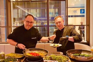 Zwei Köche stehen lächelnd an einem bunten Buffet und bereiten Gerichte mit frischem Gemüse zu
